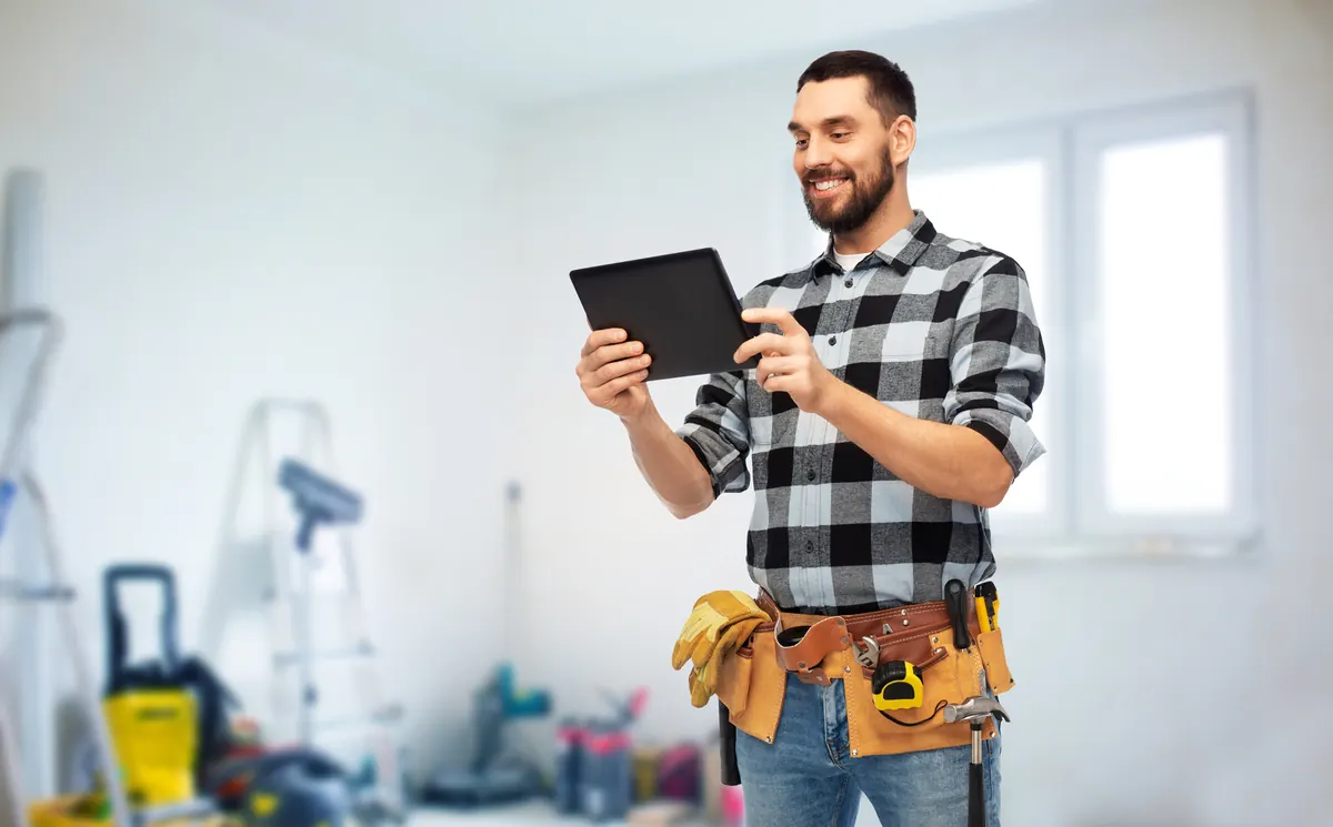 Smiling contractor in a tool belt checking a tablet on a job site